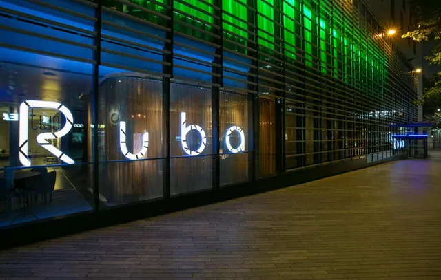 At night, an illuminated building exterior shows large lit lettering behind glass, a green-lit louvred facade, and an empty paved walkway in front.