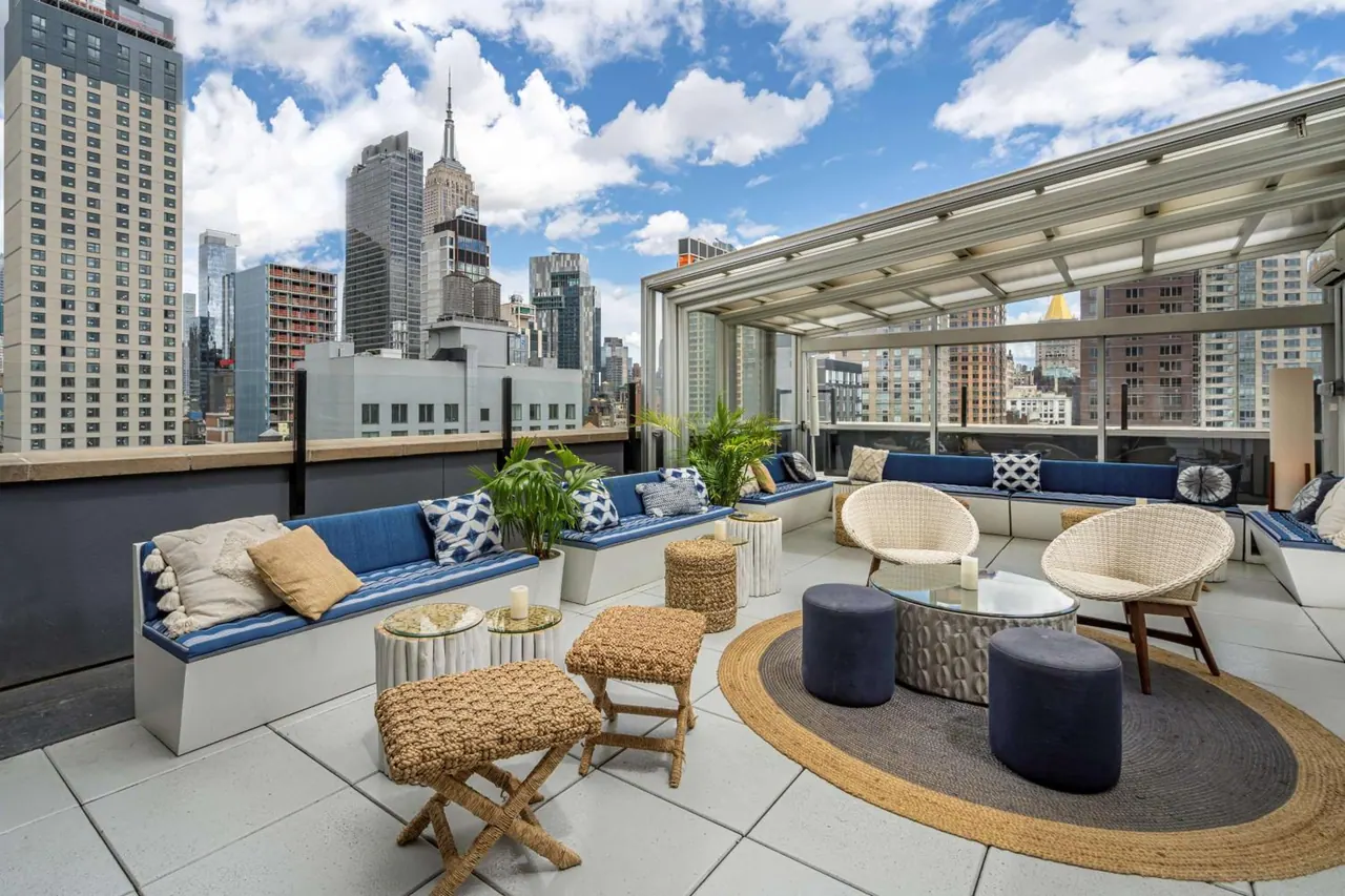 A rooftop terrace with built-in blue cushioned benches, wicker armchairs and a round rug, with the Empire State Building visible across the city skyline.