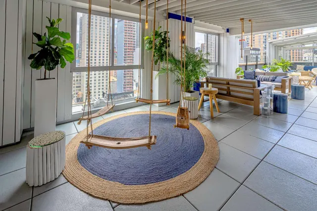A lounge area with hanging wooden swings over a round rug, potted plants, a wooden sofa and windows looking onto city buildings.
