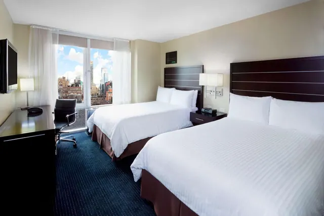 A hotel bedroom with two double beds, a desk and chair, and floor-to-ceiling windows looking out over city buildings.