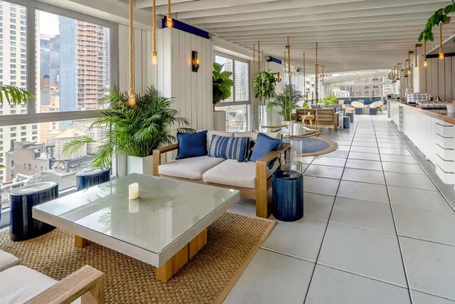 A covered rooftop lounge with blue-cushioned sofas, a glass coffee table, potted palms and a bar counter on the right, with city buildings visible through the windows on the left.