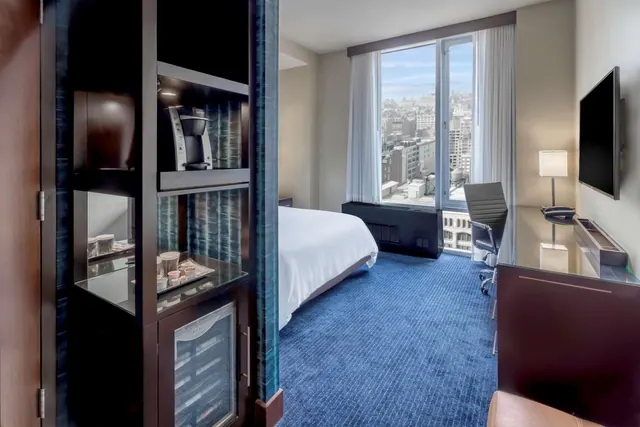 A hotel bedroom with a white king bed, a work desk with a chair, a wall-mounted television, and a window looking over city buildings, with a coffee machine and minibar in a cabinet by the entrance.