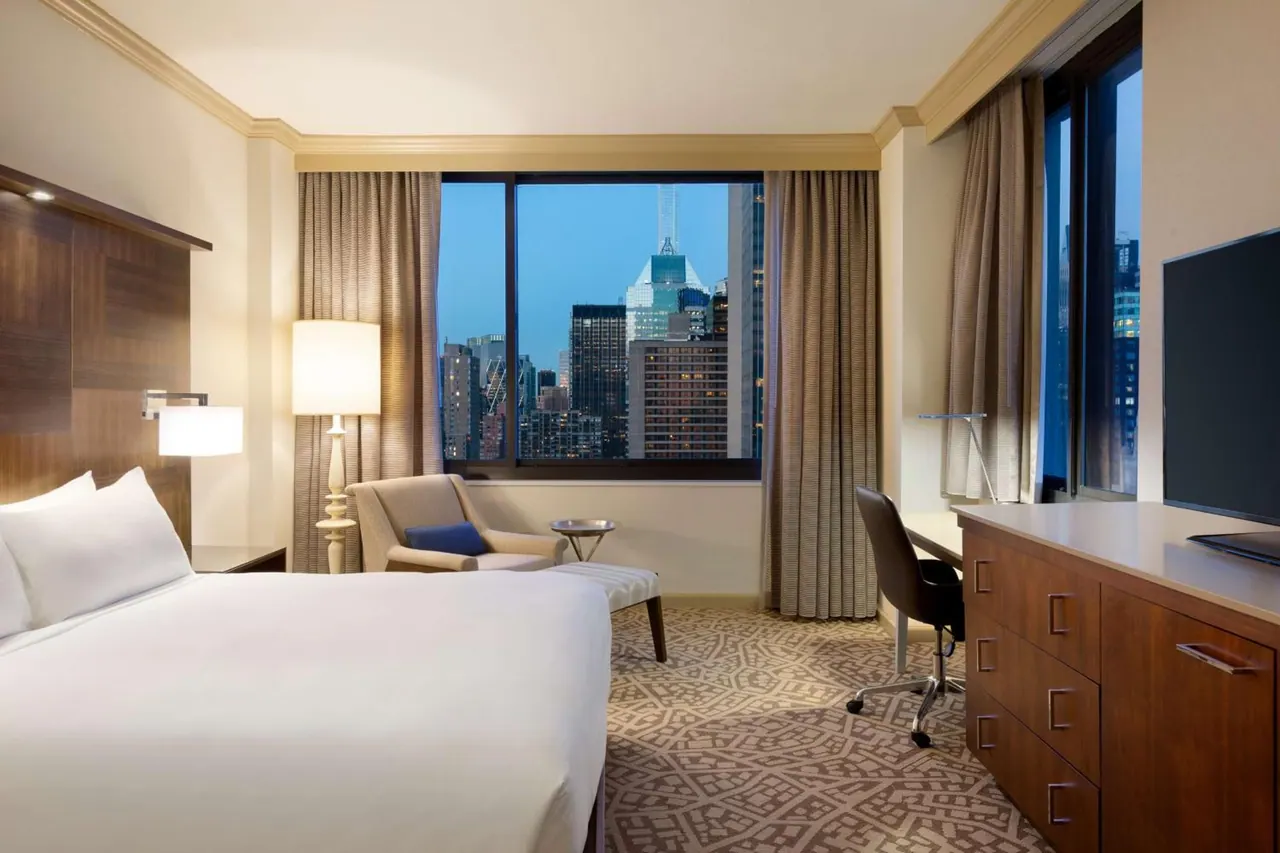 A hotel bedroom with a king bed, an armchair, a desk and a flat-screen television, with a window showing a city skyline at dusk.