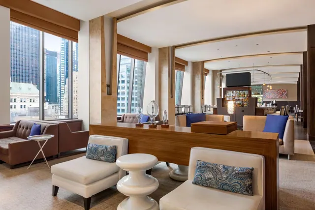 A lounge with white and brown seating, a long wooden console table, and floor-to-ceiling windows showing city high-rises.