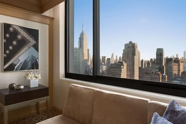 A sitting area with a brown sofa and side table beside a large window showing the Empire State Building and a midtown skyline.