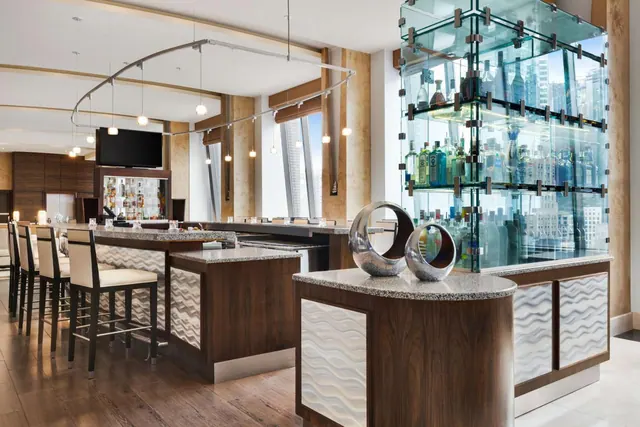 A bar area with a row of stools, a wall-mounted television, a glass-fronted bottle display and city buildings visible through the windows.
