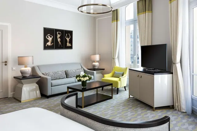 A bright sitting area has a sofa, armchair, television, decorative lighting and tall windows overlooking city buildings.