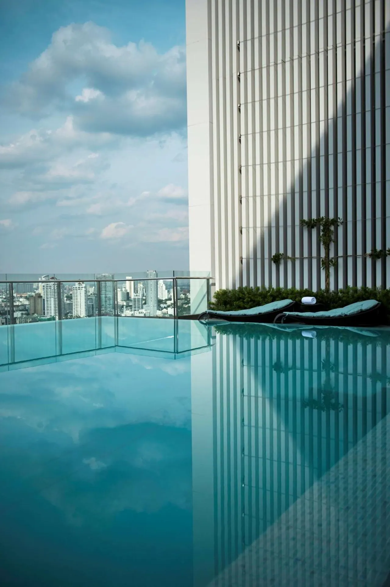 An infinity-edge rooftop pool with a glass railing overlooks a city skyline, with two sun loungers and a rolled white towel beside a tall striped building wall.