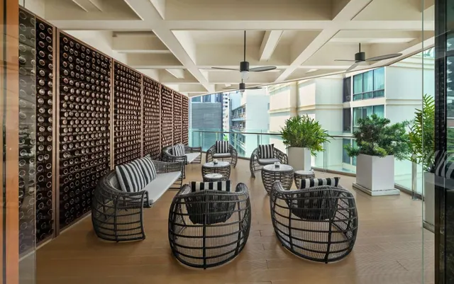 A covered terrace lounge with a wall of wine bottles, wicker armchairs with striped cushions, round side tables, ceiling fans and potted plants, with nearby buildings visible beyond the glass railing.