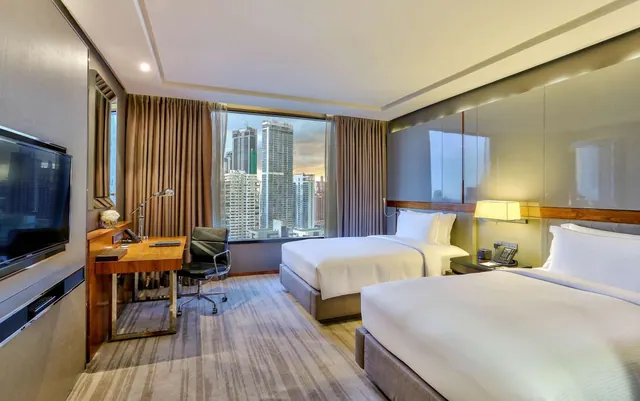 A hotel bedroom with two beds, a wooden desk and chair, a wall-mounted television, and a window showing city high-rises at dusk.