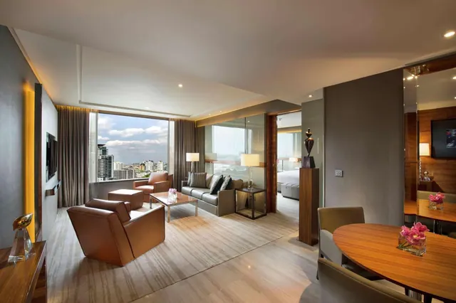 A modern living area with leather armchairs, a grey sofa, a coffee table and a wall-mounted television, with a city skyline visible through the large window and a bed visible through the doorway.