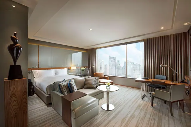 A hotel bedroom with a king bed, a grey sofa and round side table, and a wooden desk beside a large window overlooking city towers.