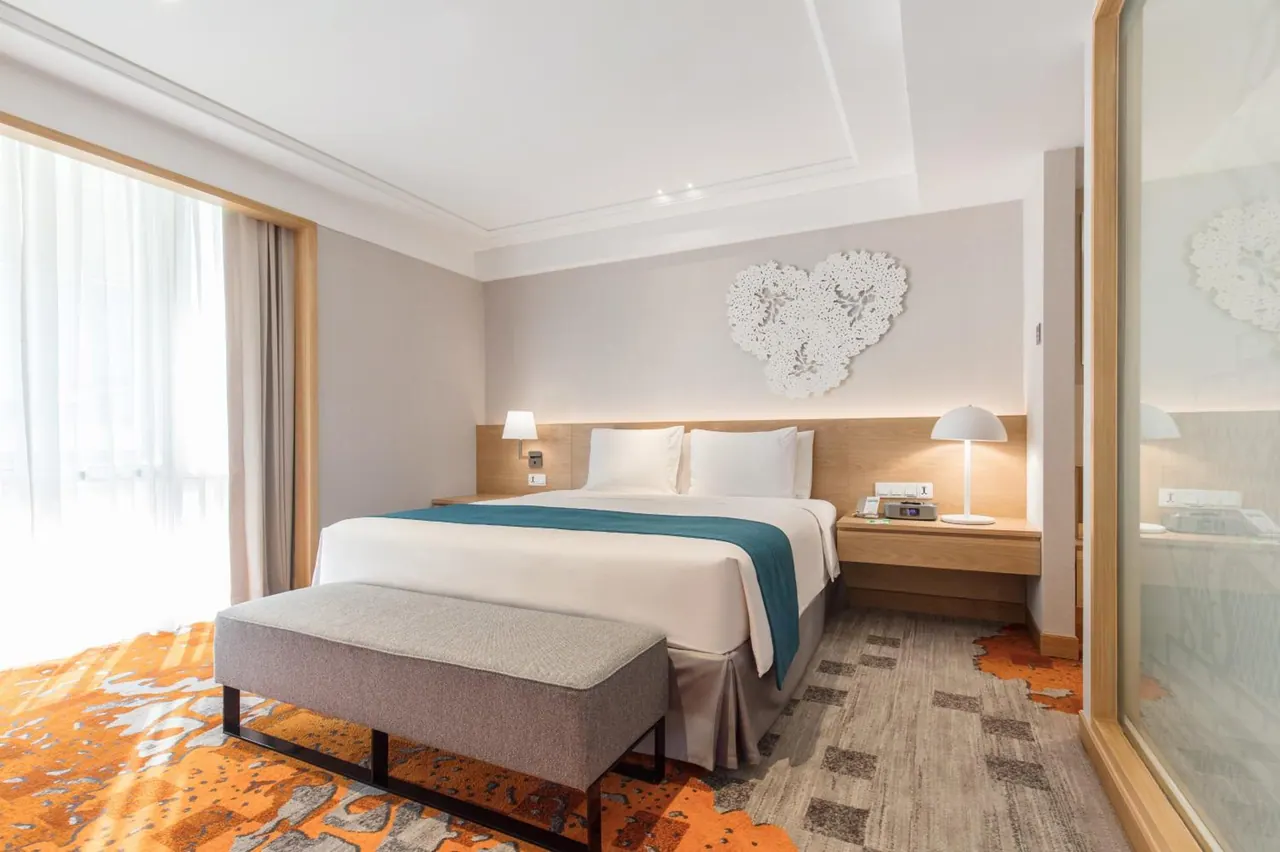 A bedroom with a made king bed with a teal runner, an upholstered bench at its foot, bedside lamps and a tall window with sheer curtains.
