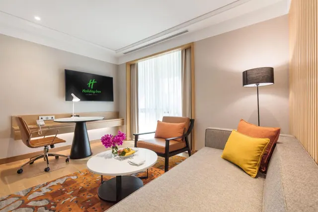A hotel room living area with a grey sofa, orange and yellow cushions, an armchair, a round white table holding fruit and a flower vase, a wall-mounted desk with a chair, and a wall-mounted television.