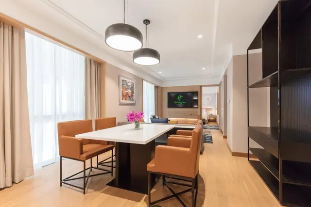 A dining table with four orange leather chairs and a vase of pink flowers sits in a suite living area with a wall-mounted TV and a bed visible through the doorway at the back.