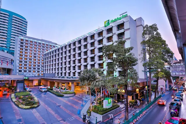 Exterior of a multi-storey Holiday Inn hotel building at dusk, with a tree-lined street, passing vehicles and a few pedestrians nearby.