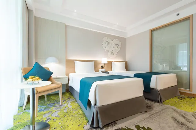 Hotel bedroom with two twin beds with teal runners, a teal-cushioned armchair, and a small round table holding a plate of fruit.