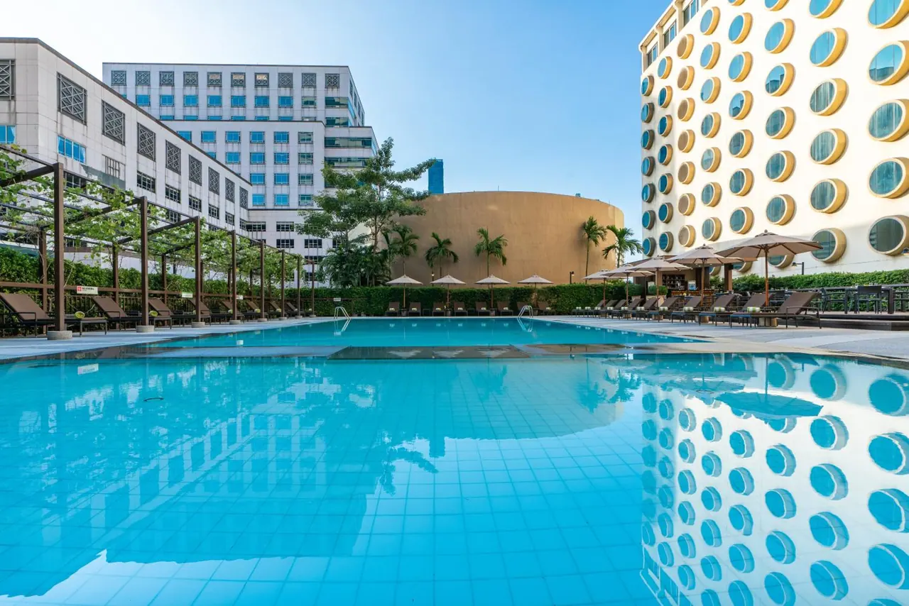 A large outdoor swimming pool with blue tiles, surrounded by sun loungers and parasols, with hotel buildings with round windows in the background.