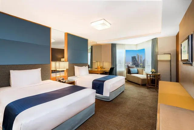 Twin-bed hotel room with a wall-mounted television, a lounge chair by a window showing city towers, and a desk in the background.