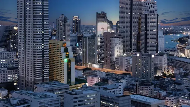 Twilight view over a dense Bangkok skyline of high-rise towers and low-rise buildings, with a river visible on the right.