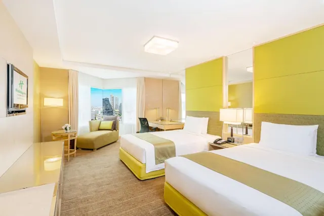 A hotel bedroom with two beds against yellow-green headboard walls, a seating chair, a desk, a television and a window showing city buildings.