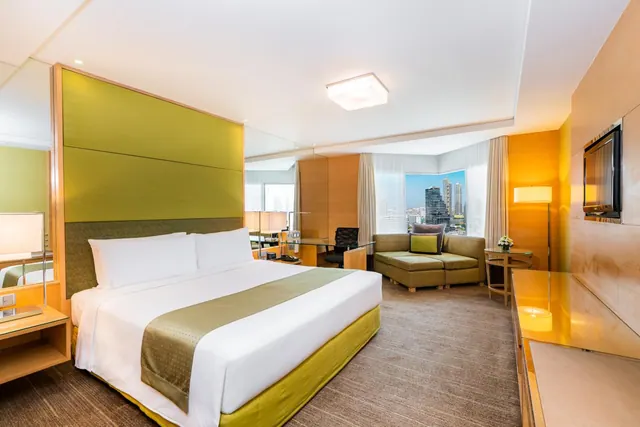 A hotel room with a king bed under a green headboard panel, a sofa and ottoman, a work desk, and a window overlooking city towers.