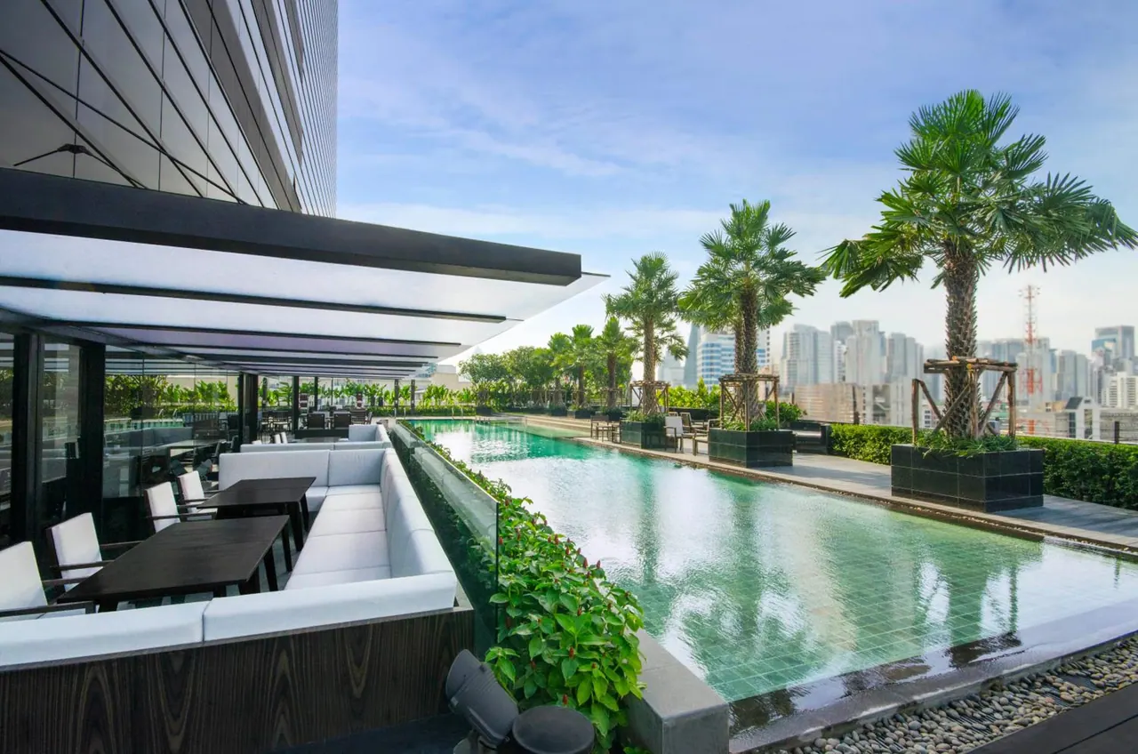 A long rooftop swimming pool with palm trees and white lounge seating beside it, with city towers visible in the background under a blue sky.