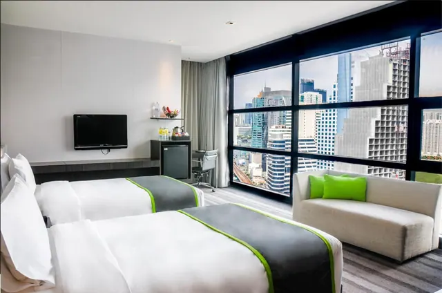 A hotel bedroom with two beds, a sofa, a wall-mounted television and floor-to-ceiling windows looking out over high-rise buildings.