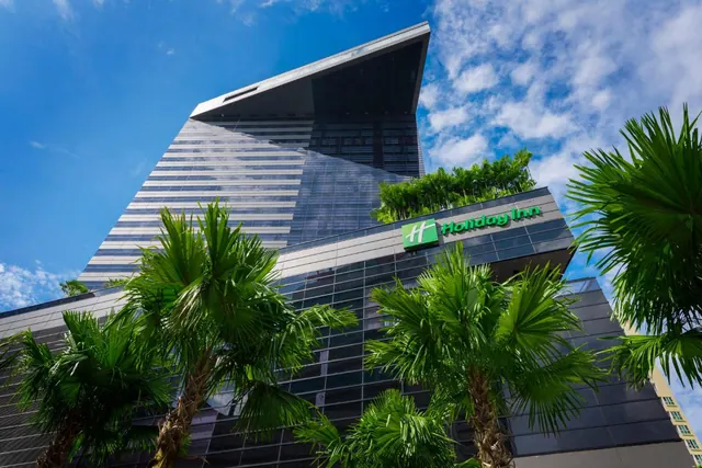 Low-angle view of a tall glass-and-metal hotel tower with a Holiday Inn sign, framed by palm trees under a blue sky.