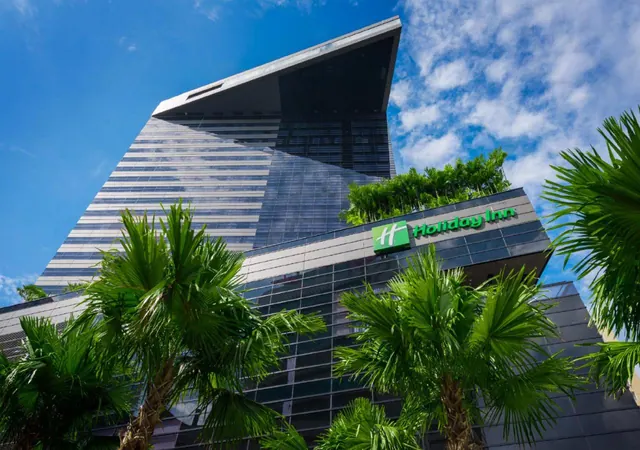 Low-angle view of a modern glass hotel tower with a Holiday Inn sign, framed by palm trees under a blue sky with clouds.