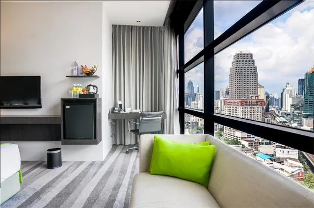 A hotel room with a grey sofa and green cushion, a wall-mounted television, a minibar, a desk and chair, and floor-to-ceiling windows overlooking city high-rises.