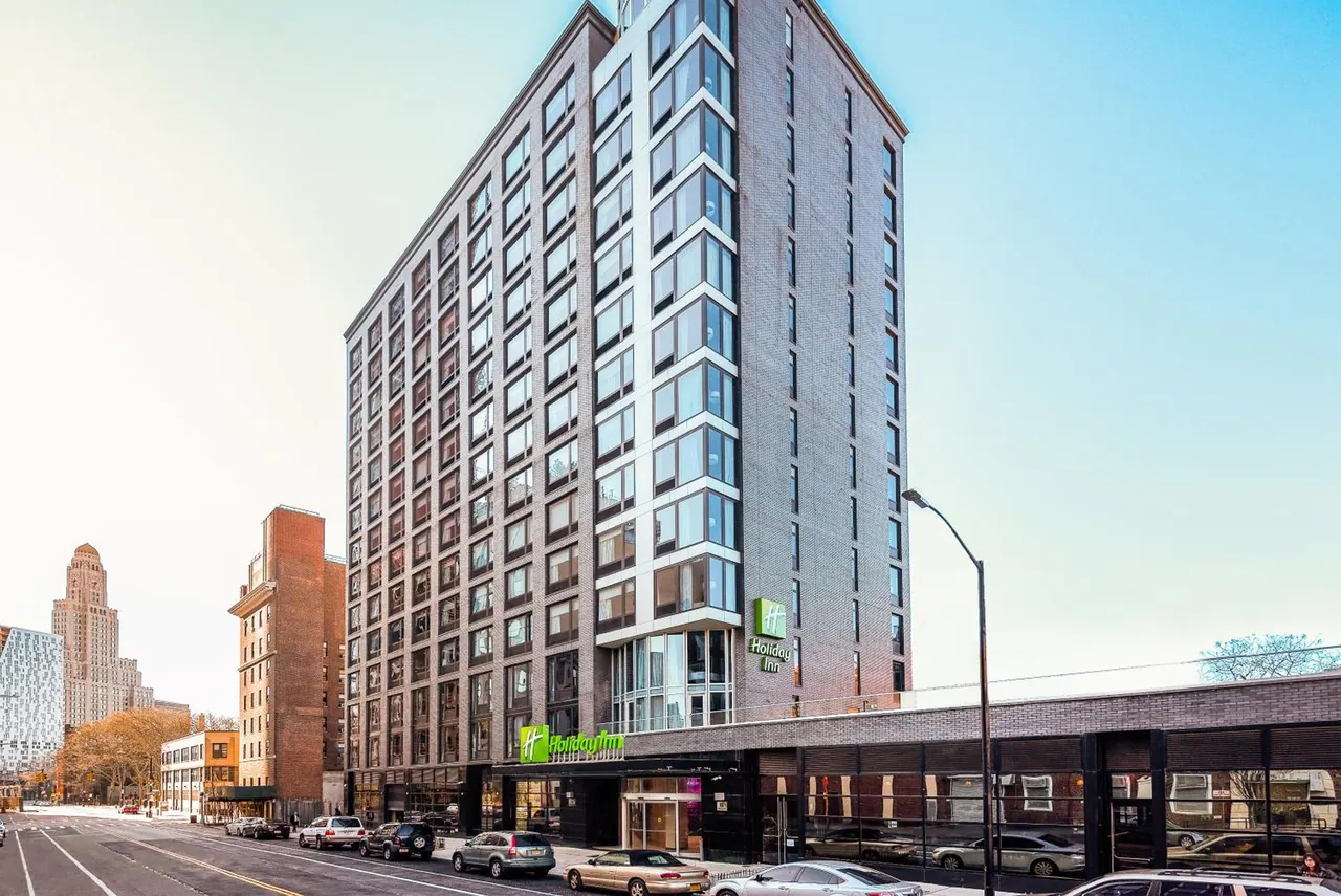 A tall brick Holiday Inn building with a green sign rises above a street lined with parked cars.