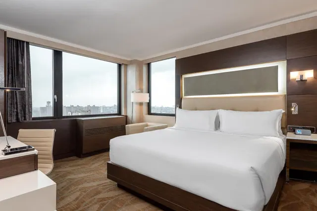 A king bed with white linens sits across from a desk and armchair beside large windows that look out over city buildings.