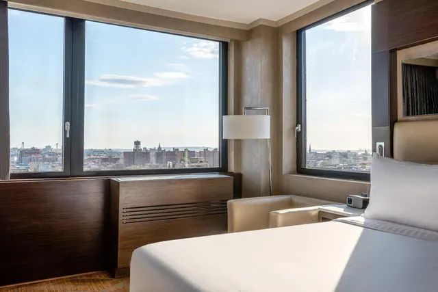 A hotel bedroom with a made bed, a cream armchair and a floor lamp, with large windows looking out over a city skyline.