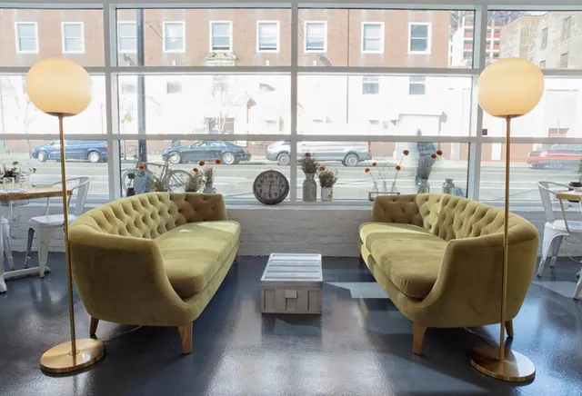 Two olive velvet tufted sofas face each other in a lounge with floor lamps and a large window looking onto a street with parked cars and brick buildings.