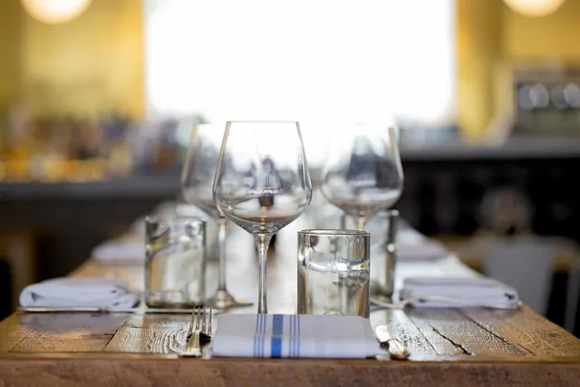 A set restaurant table with wine glasses, water glasses, folded napkins and cutlery on a wooden table, with a bright window and a bar blurred in the background.