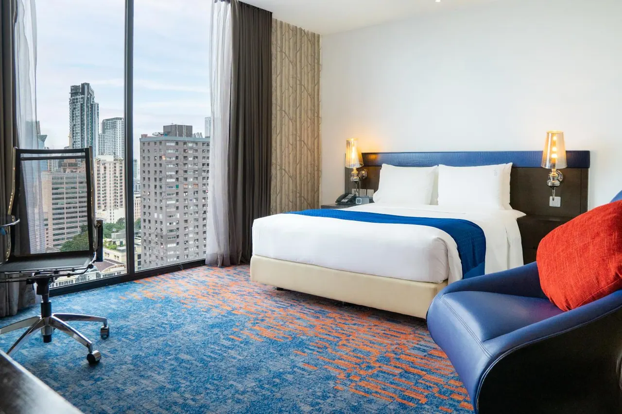 A hotel bedroom with a king bed, a blue armchair, a desk and chair, and floor-to-ceiling windows overlooking high-rise buildings.