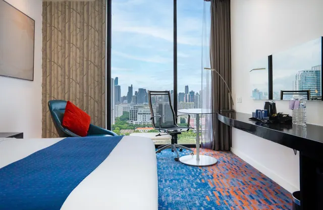 A hotel bedroom with a double bed, an armchair, a desk with a kettle and water bottles, and floor-to-ceiling windows overlooking a city skyline.