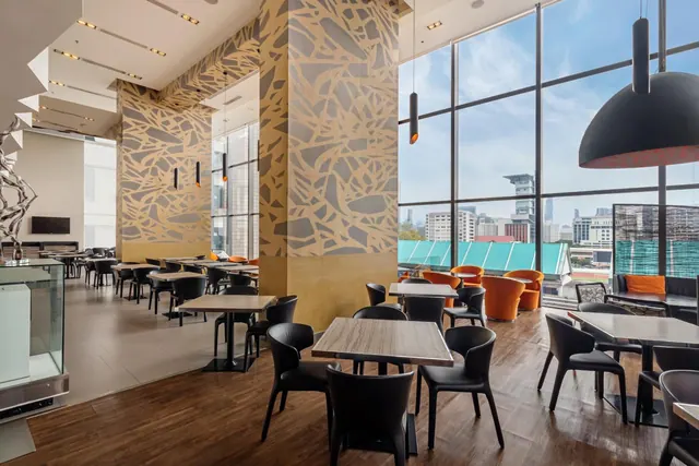 A dining room with black chairs and wooden tables, orange armchairs, pendant lights, and floor-to-ceiling windows looking out over city buildings.
