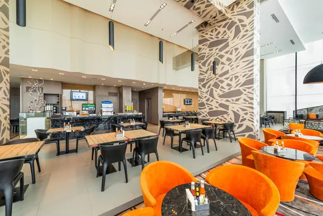 A restaurant dining area with wooden-top tables, black chairs, orange armchairs, patterned columns and a buffet counter in the background.