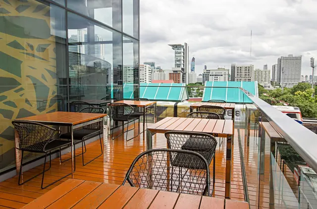 Outdoor terrace with wooden tables and woven chairs, a glass balustrade, and a view over city high-rise buildings under a cloudy sky.