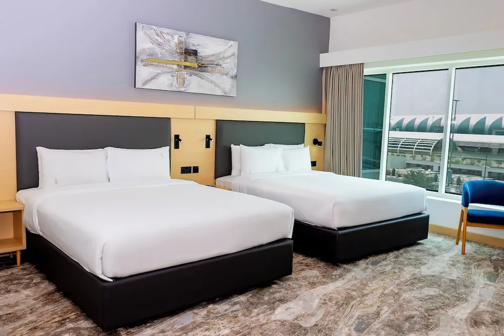 A hotel bedroom with two double beds made up with white bedding, a painting on the wall above them, and a large window looking out onto a building with a curved roof.