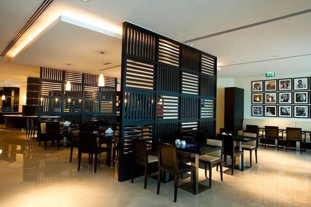 A restaurant dining room with dark slatted wooden dividers, tables with dark upholstered chairs, a built-in banquette, and framed black and white photographs on the wall.