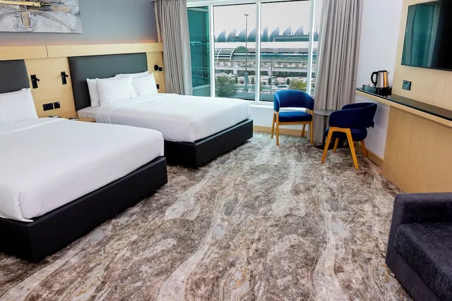 A hotel bedroom with two double beds with white bedding, blue armchairs by a window looking out onto an airport terminal and a road, a wall-mounted television, and a kettle on the side unit.