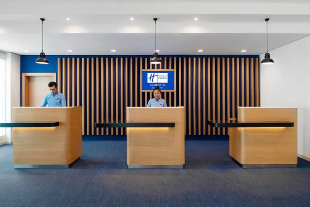 Two staff in blue uniforms stand behind three wooden reception desks in a lobby with a slatted wood wall and a Holiday Inn Express sign.