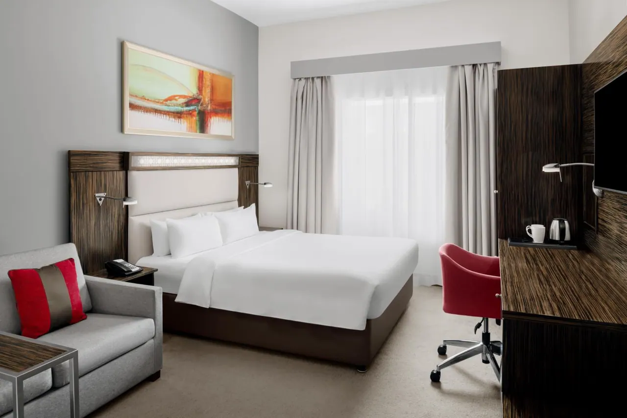 A hotel bedroom with a king bed, a grey armchair with a red cushion, a red desk chair, a wall-mounted television and a kettle on a dark wood cabinet.
