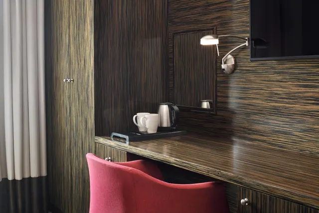 A wood-veneered desk with a red chair, a tray holding two mugs and a kettle, a wall lamp and a wall-mounted television in a hotel room.