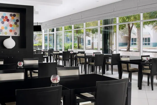 A dining room with black tables and chairs with cream seat pads, set in front of large windows looking out onto trees and a white building.