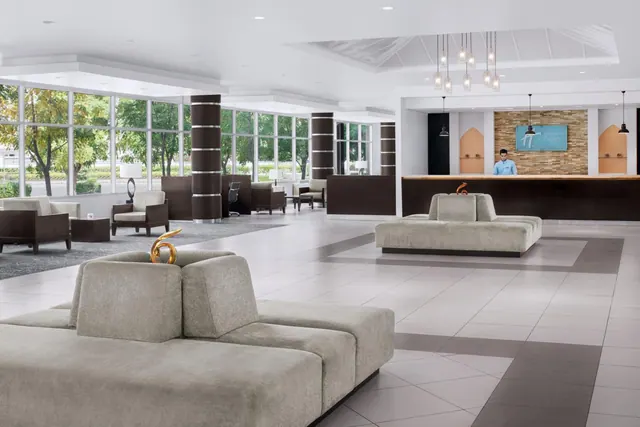 A bright modern hotel lobby with grey sofas, armchairs, a reception desk with one staff member, and large windows looking out onto trees.
