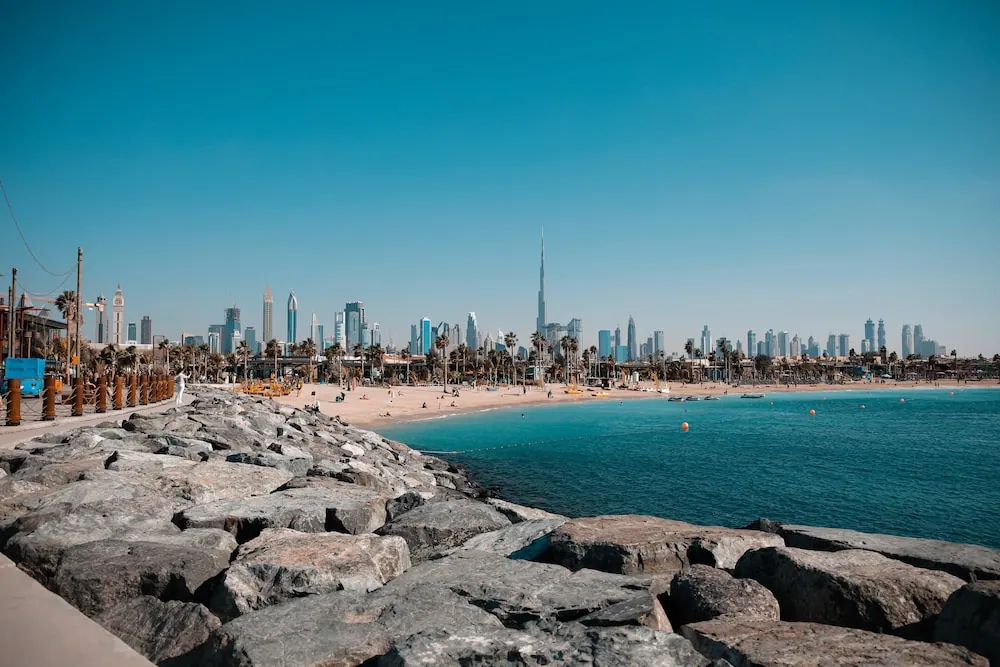 A rocky breakwater in the foreground leads to a sandy beach, turquoise sea and a city skyline with the Burj Khalifa under a clear blue sky.